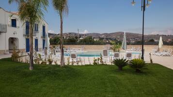 Zona de piscina con sillones de descanso, una palmera y un edificio con puertas y ventanas azules.