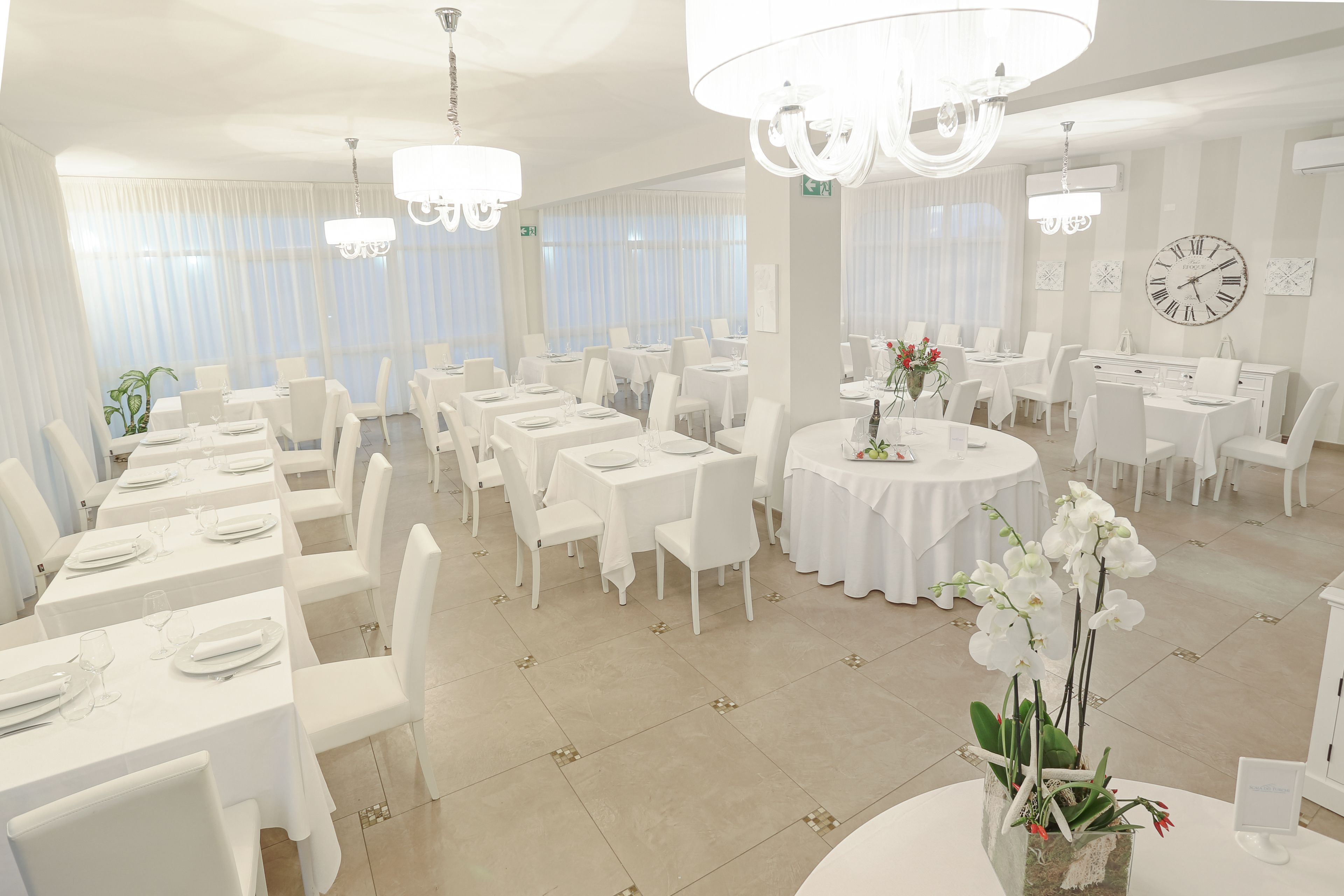 Un comedor amplio con varias mesas y sillas, ventanas grandes con cortinas translúcidas y un candelabro.