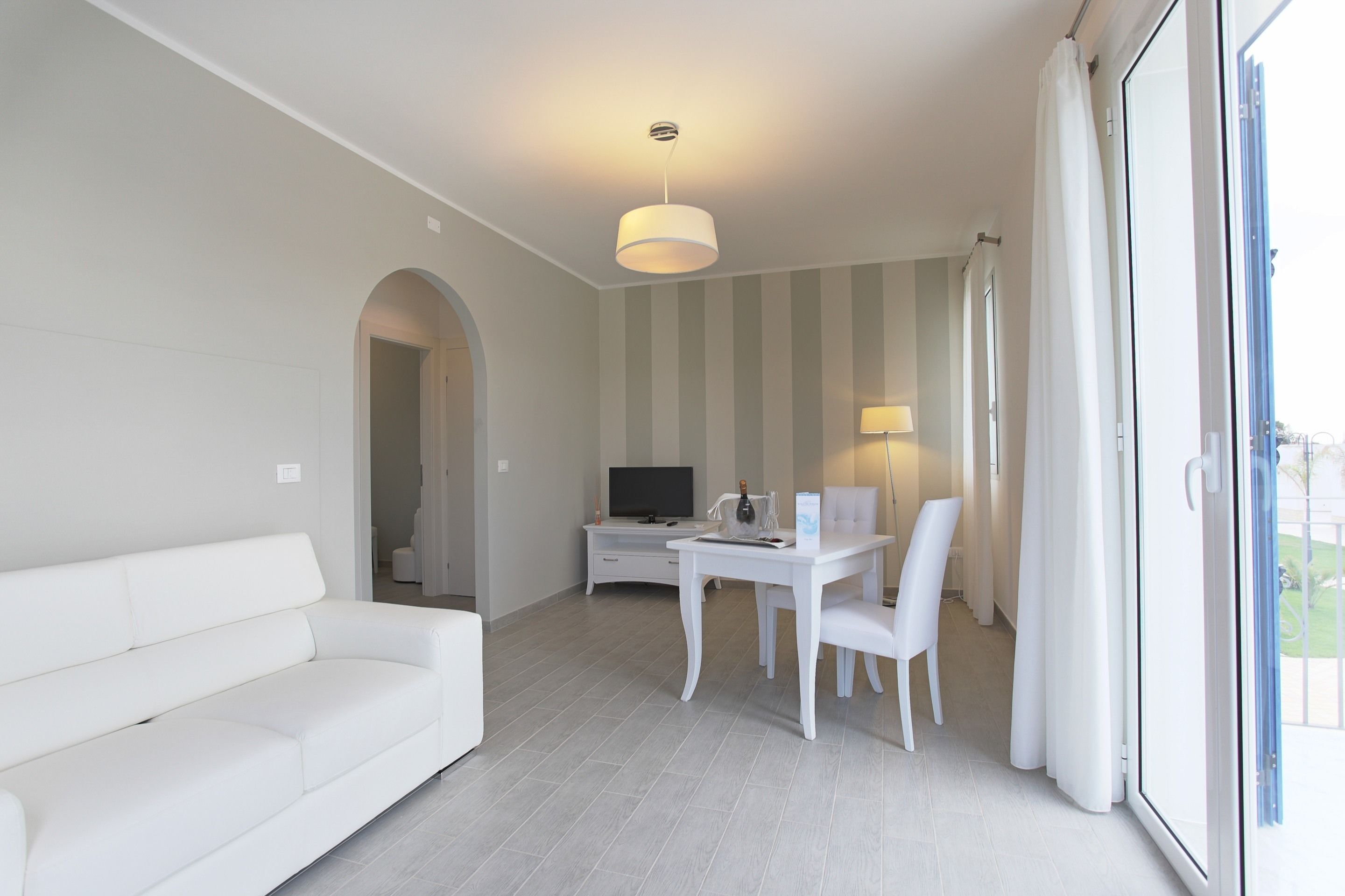 A modern living room with a white sofa, a dining table with chairs, a TV, and a large window with curtains.