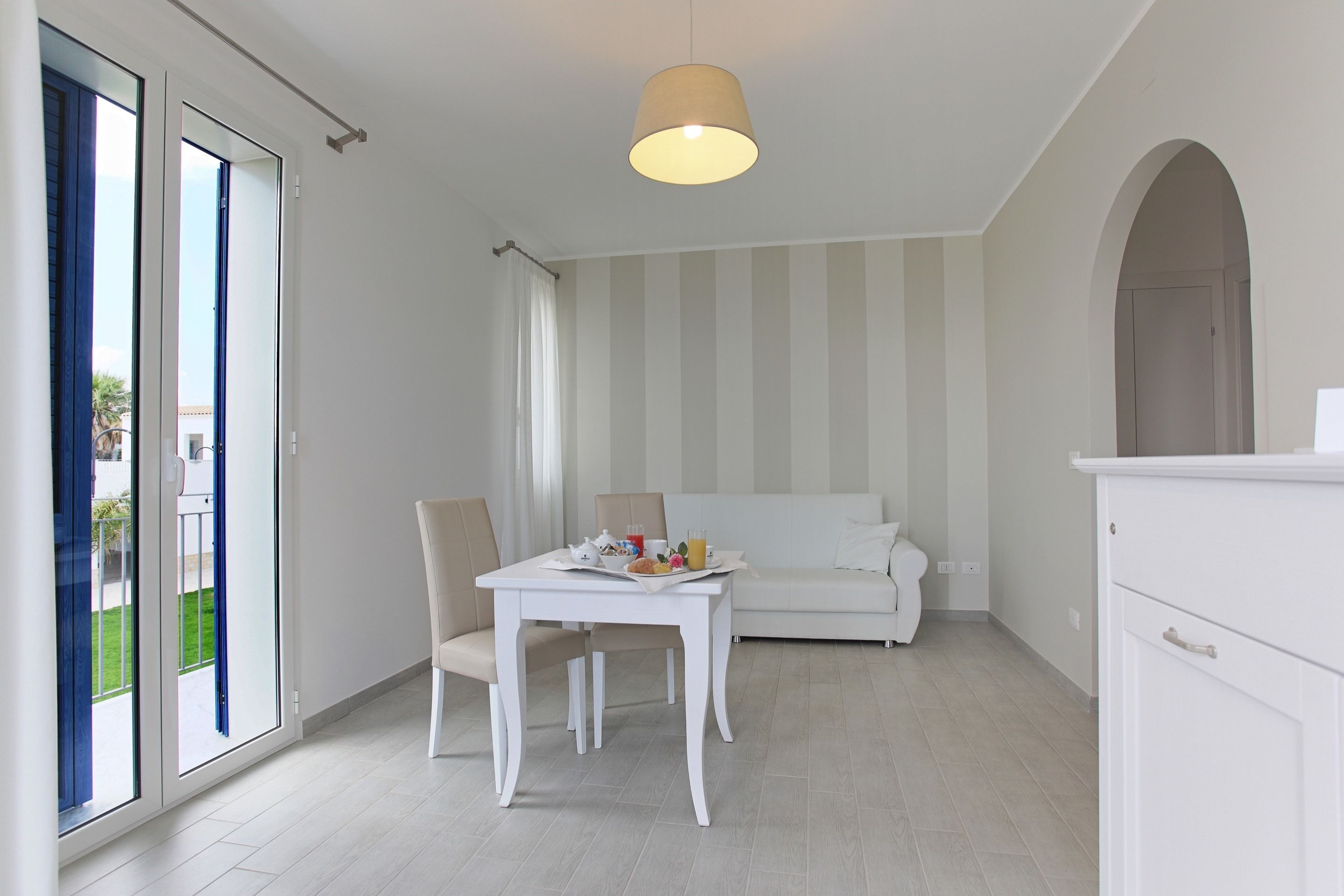 A modern, white interior with a dining table set for breakfast, a sofa, and a kitchen area.
