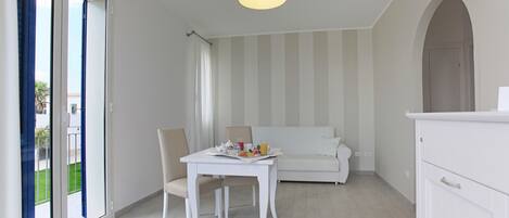 A modern, white interior with a dining table set for breakfast, a sofa, and a kitchen area.