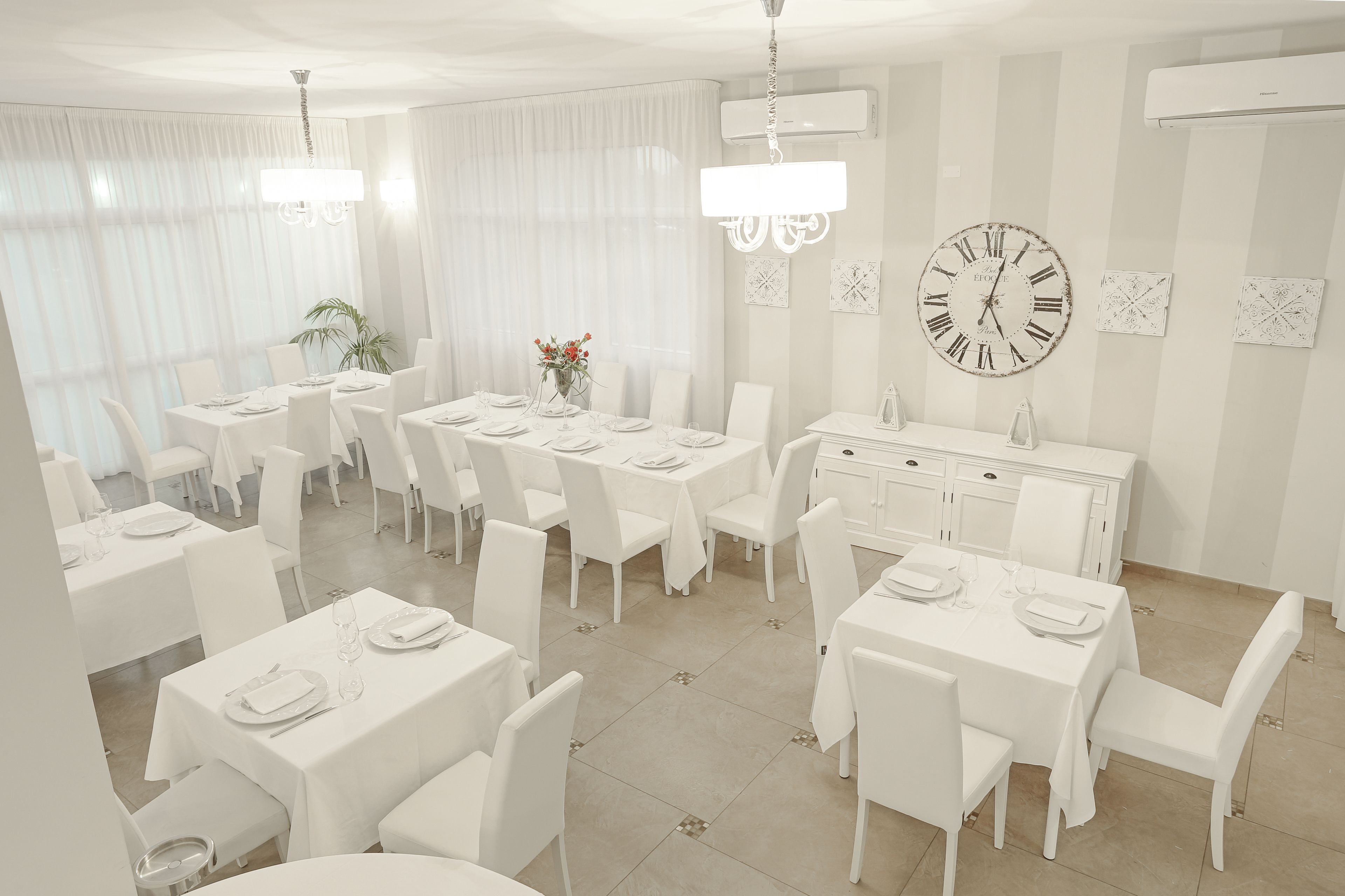 Un espace repas avec des tables et des chaises blanches, une grande horloge murale et un lustre.