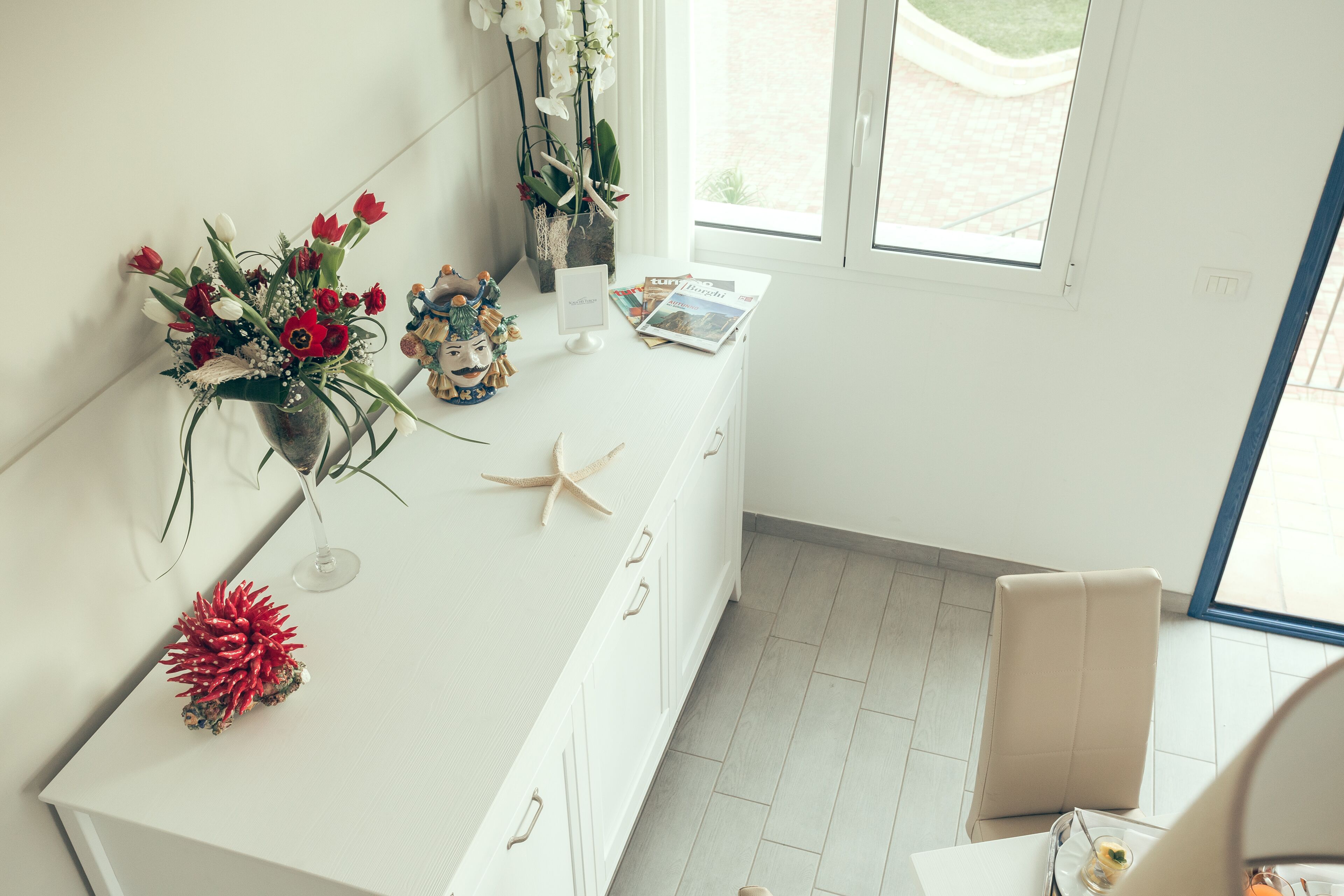 Una mesa de comedor blanca con un jarrón de flores rojas y blancas, un adorno de estrella de mar y una pequeña estatua. Se ve una ventana con vistas a un jardín al fondo.