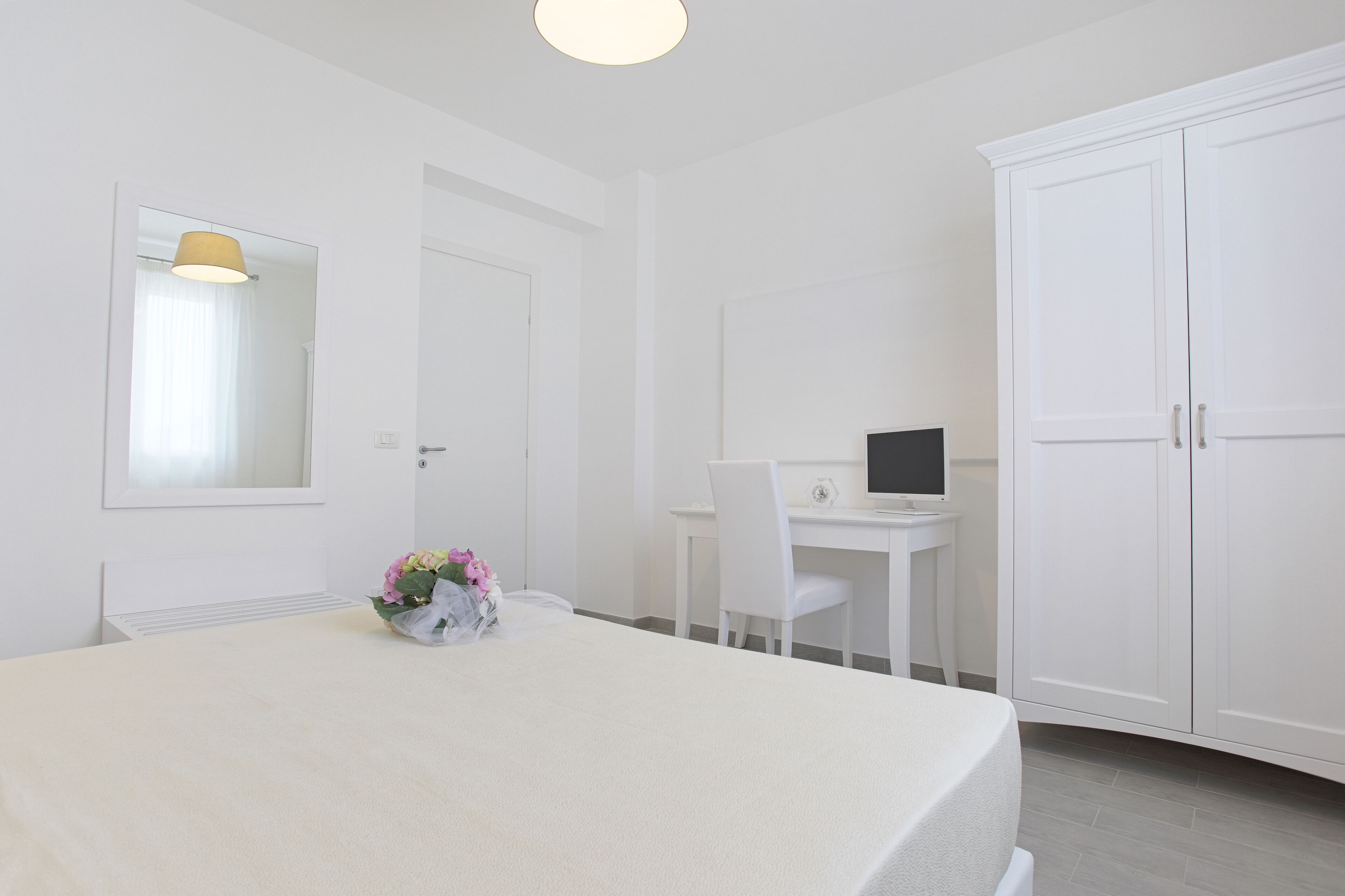Een moderne, witte slaapkamer met een bed, een bureau met een laptop en een kledingkast.