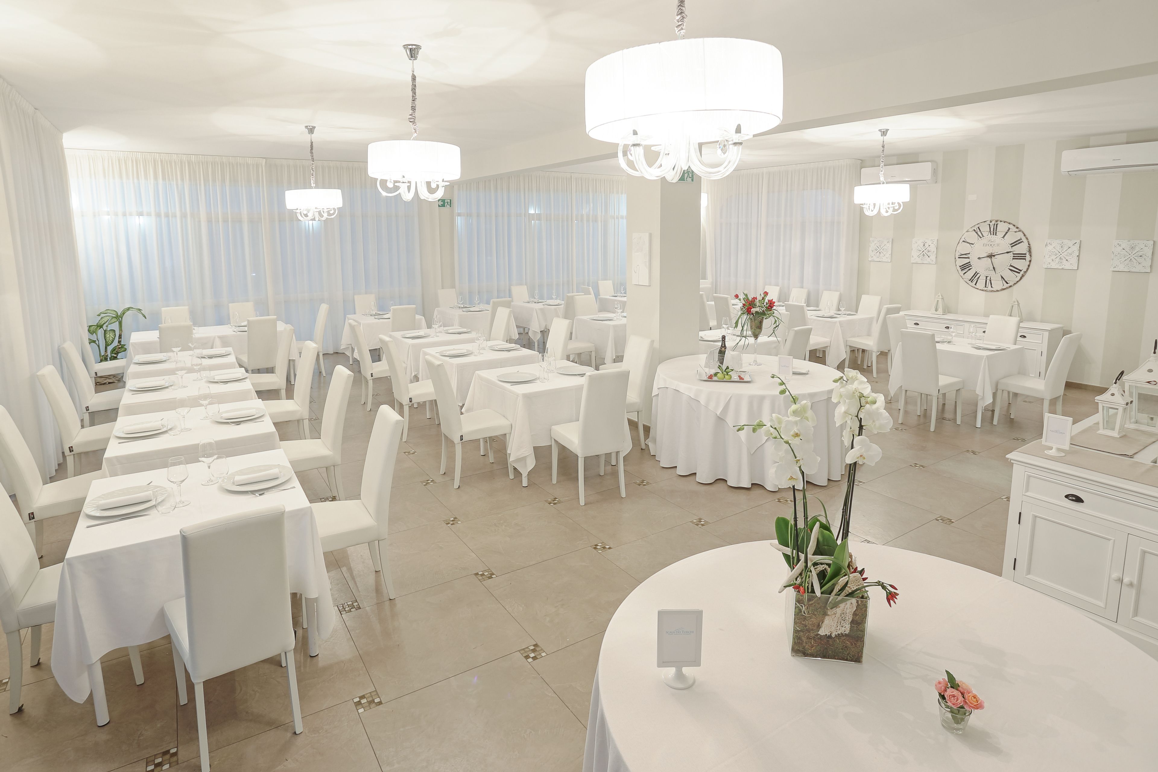 Un vaste espace dédié à la salle à manger, avec plusieurs tables rondes, des chaises blanches et des lustres.
