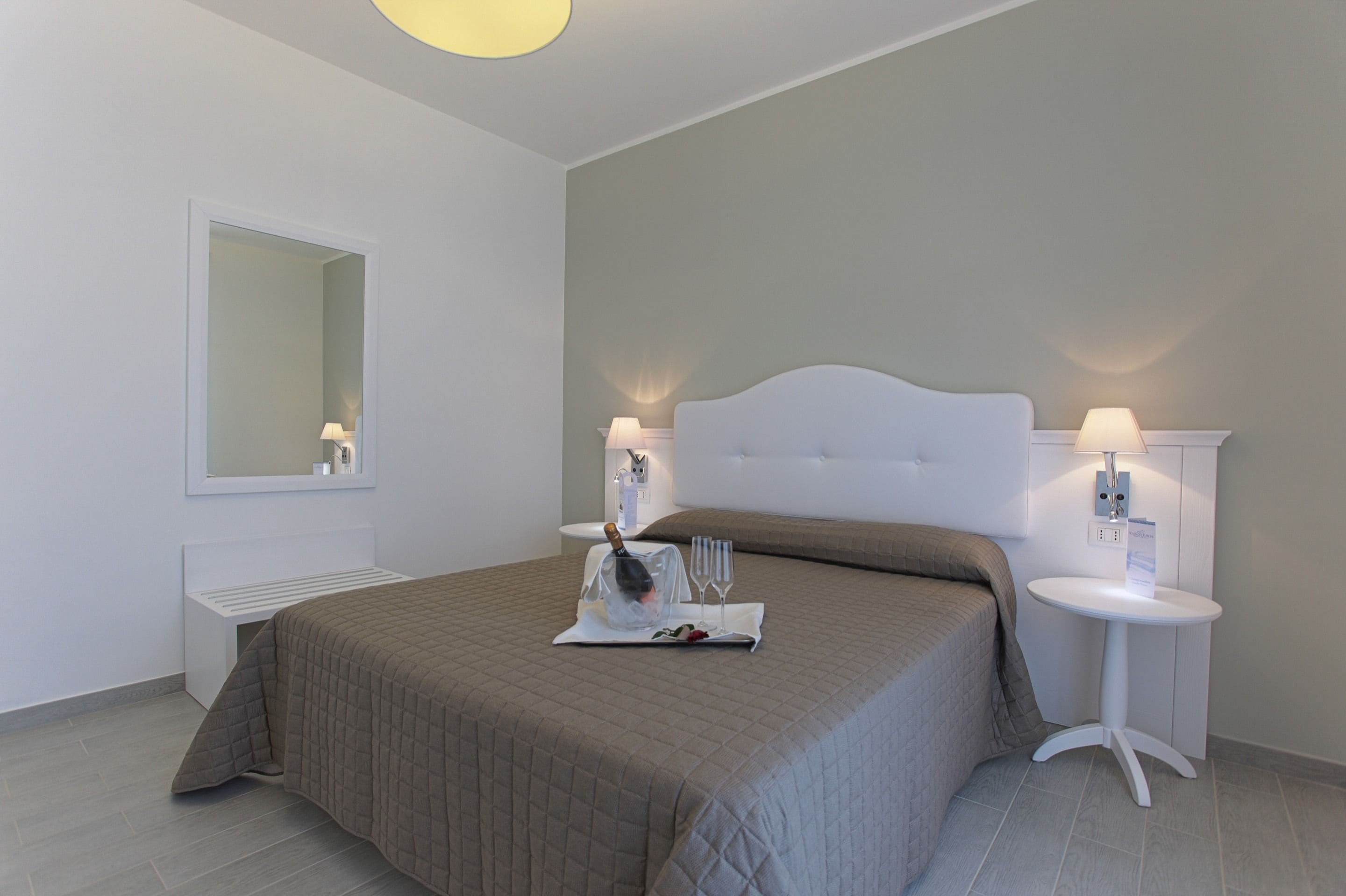 A hotel room with a large bed, two bedside tables with lamps, a small table with a bottle and glasses, and a mirror on the wall.