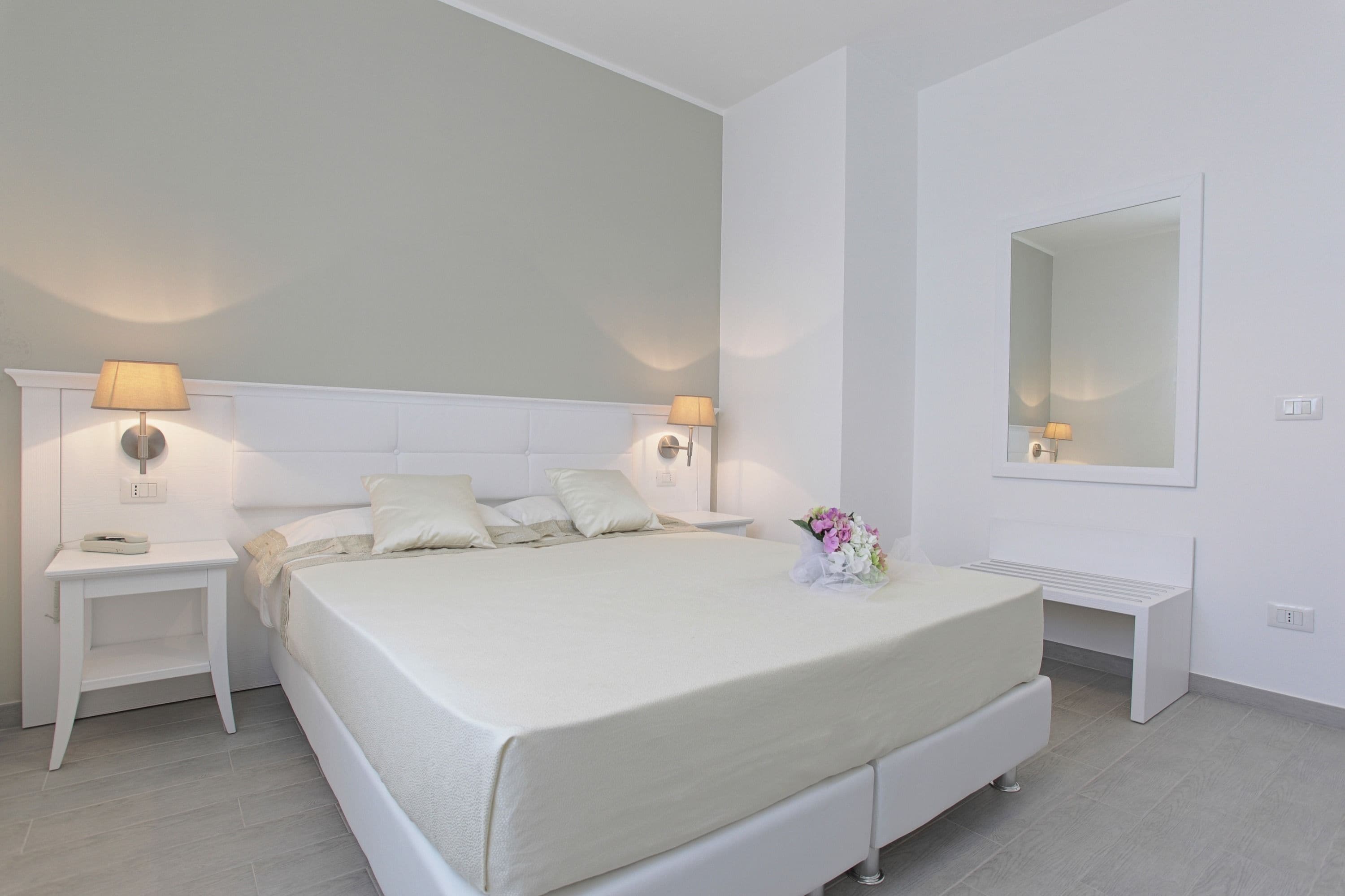 A modern bedroom with a large bed, bedside lamps, a nightstand, a bench, and a mirror.