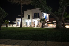 A two-story building with a white facade, blue shutters, and a balcony. There are outdoor tables and chairs arranged in the front yard. - Scala Dei Turchi Resort (Realmonte)