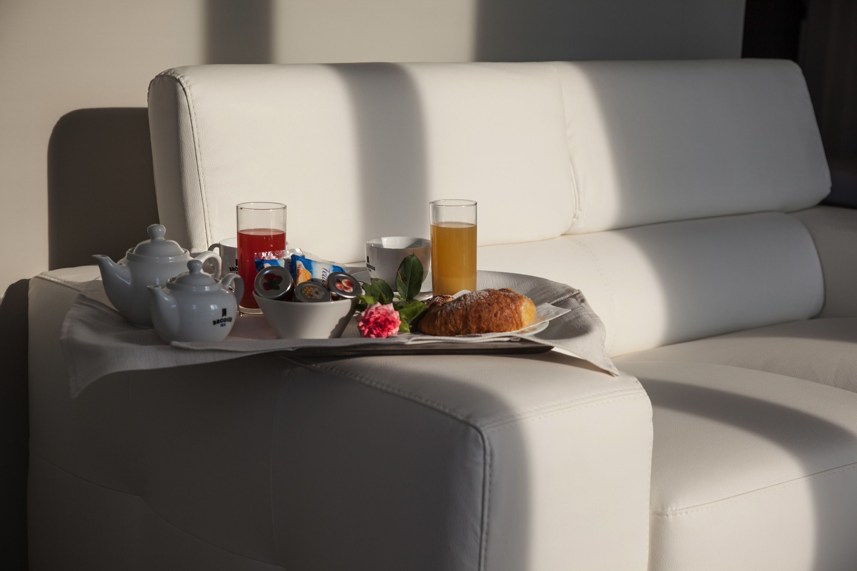 Frühstücksbrett mit Teekanne, Tassen, einem Croissant und einer Blume auf einer weißen Ledersofa.