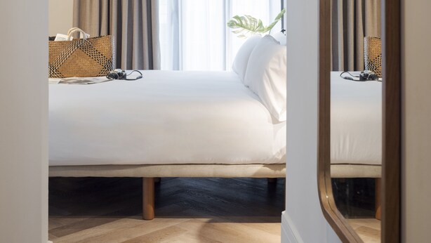 Une chambre d’hôtel avec un lit, une table de chevet, une lampe, une plante et une fenêtre avec des rideaux.