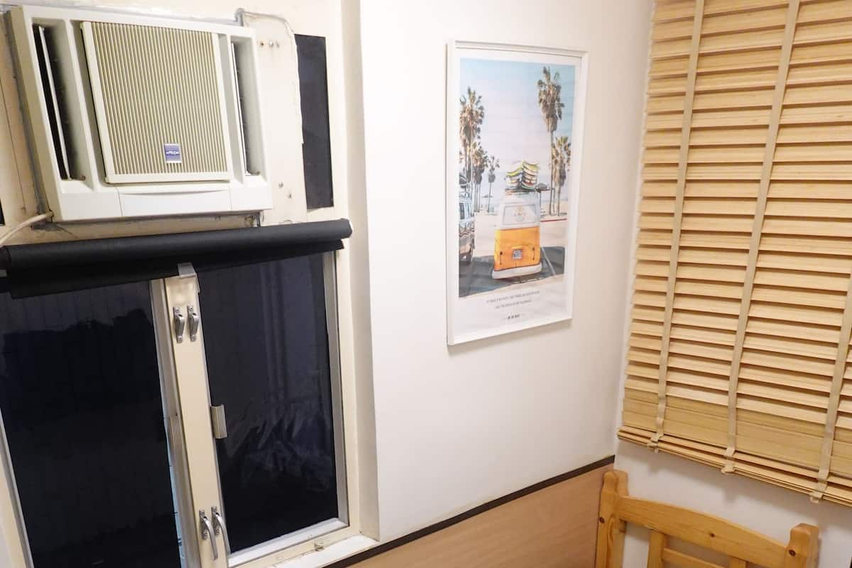 A small, single-bed room with a window, a wall-mounted air conditioner, and a framed picture.