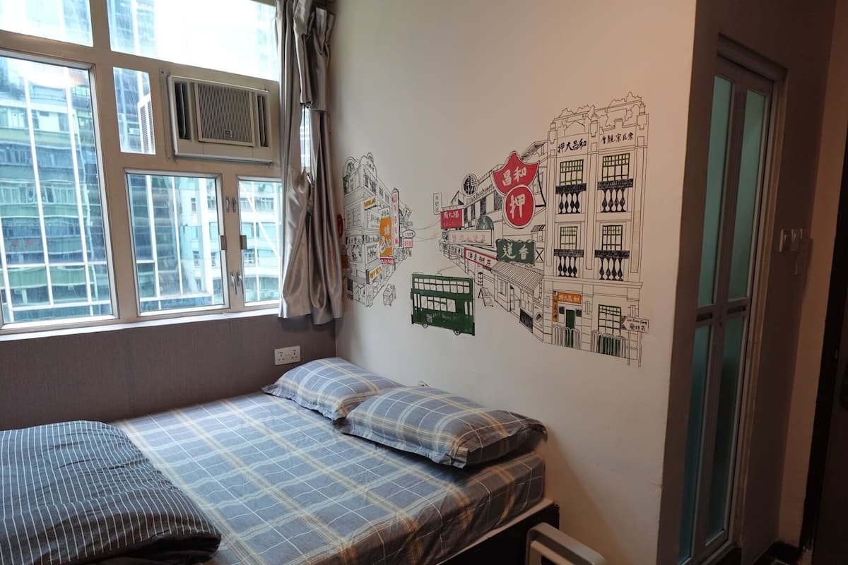 A small, single-bed room with a window, a radiator, and a wall decorated with a cityscape mural.