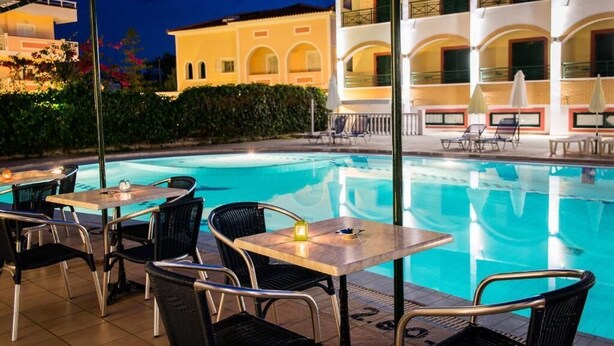 An outdoor pool area with tables and chairs, a building with balconies, and a hedge in the background. - Club Zante Plaza (Zakynthos)
