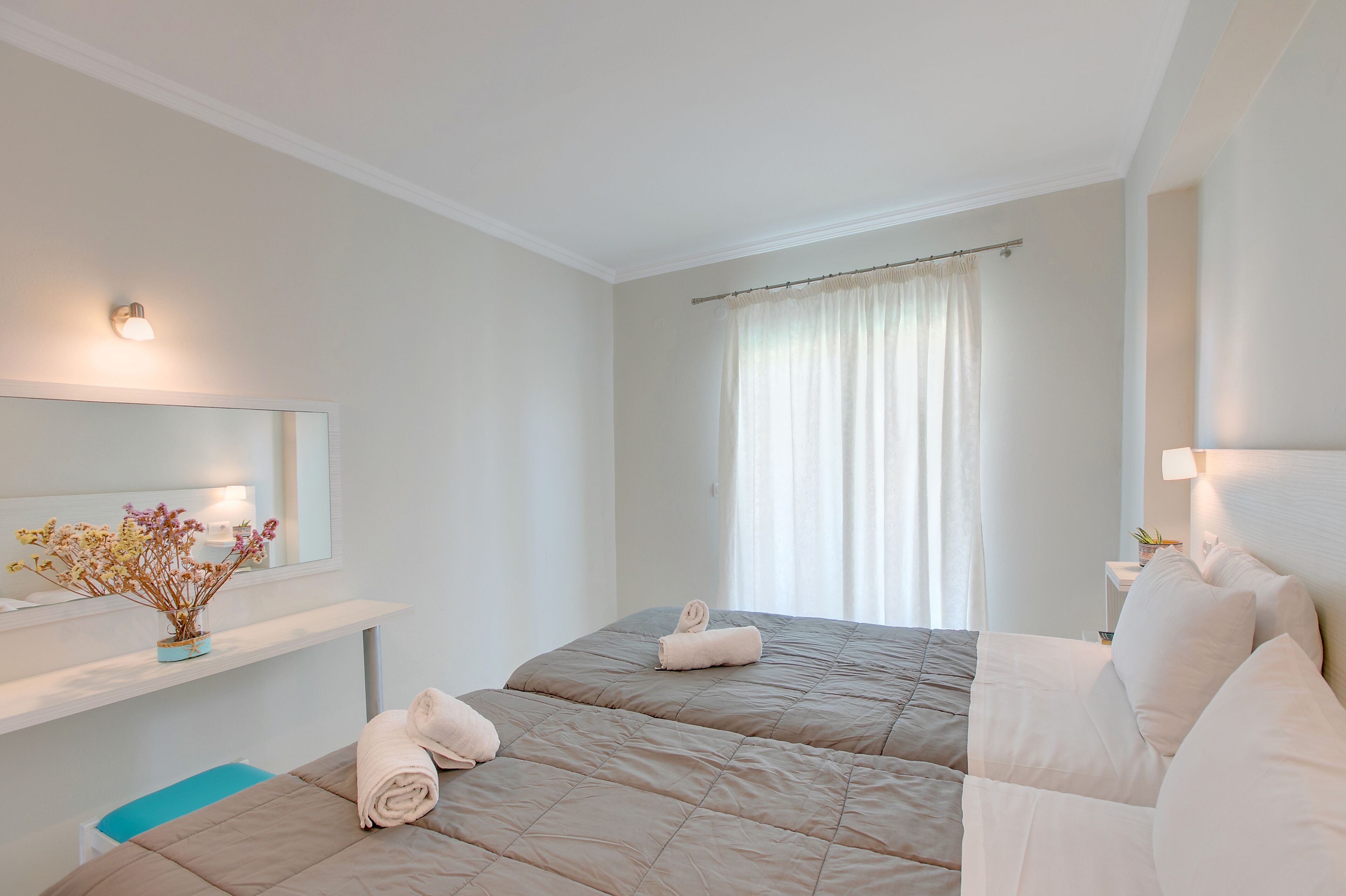 A hotel room with two beds, a bedside table with a lamp, a mirror, and a window with curtains.