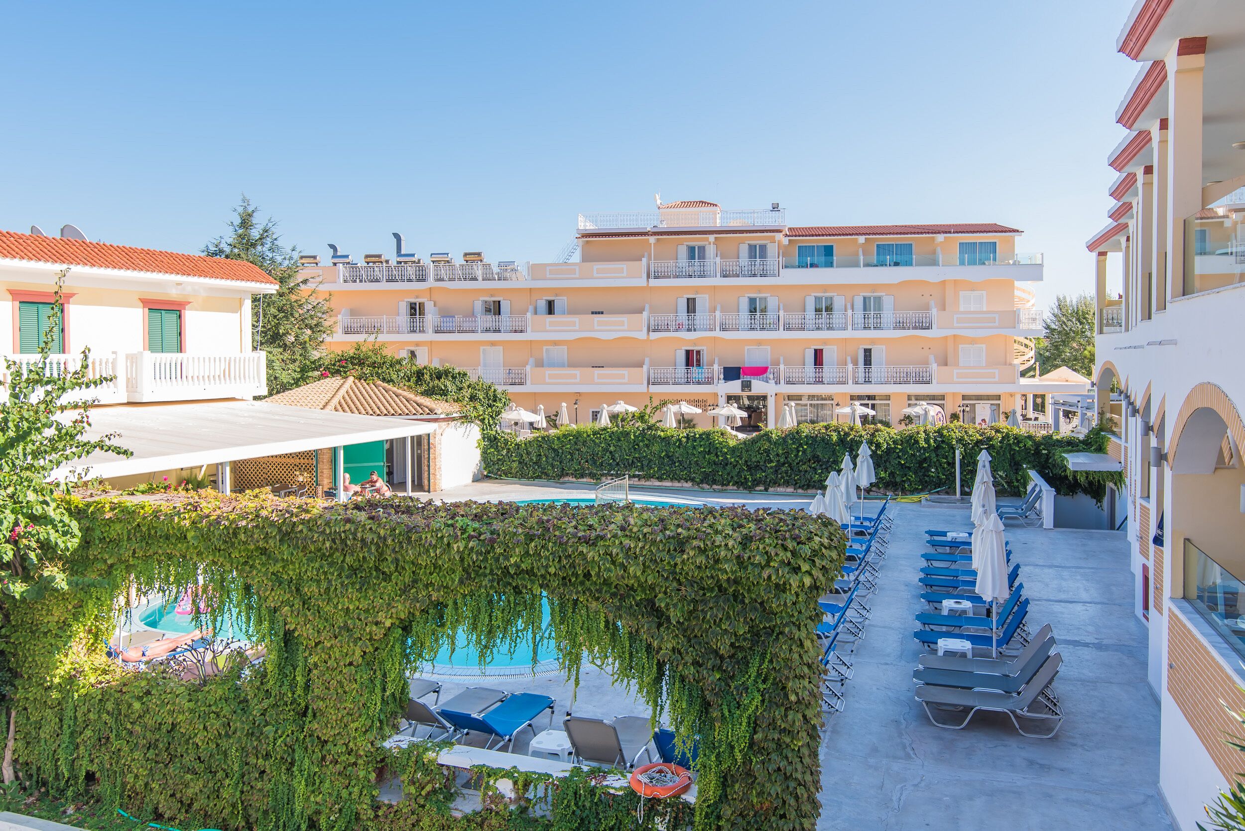 Un hôtel doté d’une piscine entourée de chaises longues et de parasols, d’un espace couvert avec une table et des chaises, et d’un bâtiment avec des balcons et un toit recouvert de tuiles rouges.