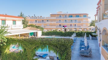 Un hôtel doté d’une piscine entourée de chaises longues et de parasols, d’un espace couvert avec une table et des chaises, et d’un bâtiment avec des balcons et un toit recouvert de tuiles rouges.