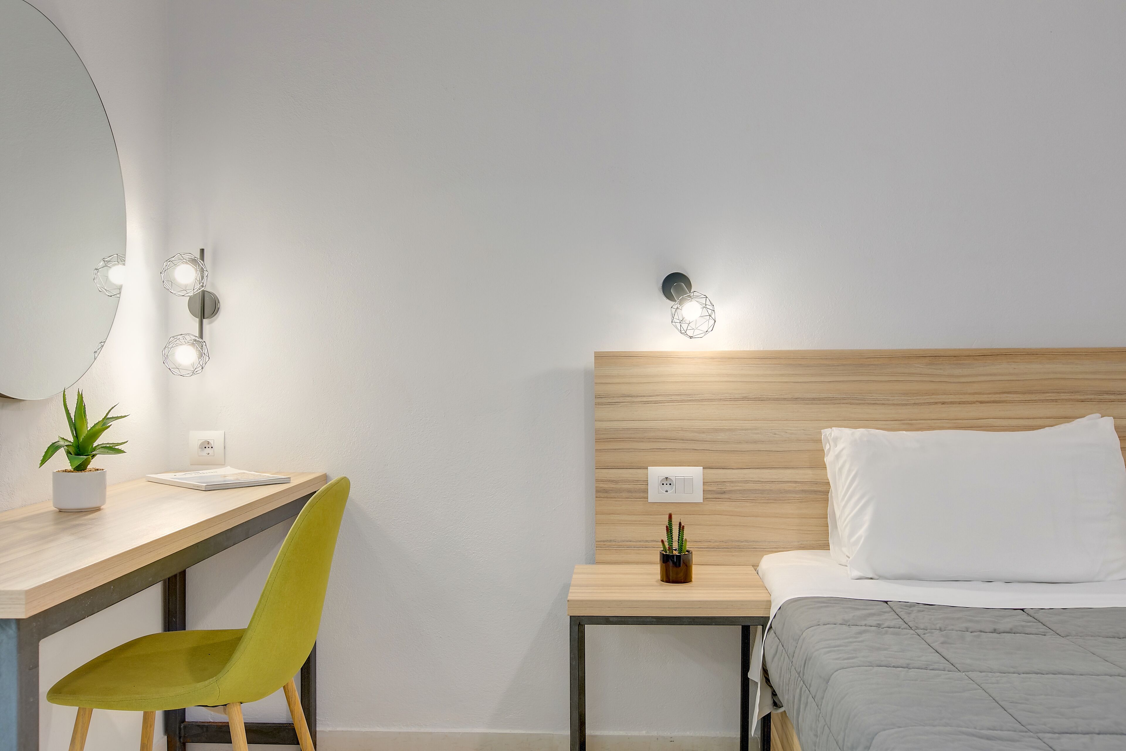 Habitación de hotel moderna con tablero de cabecera de madera, un escritorio con silla, una mesita de noche con planta y una cama con colcha gris.
