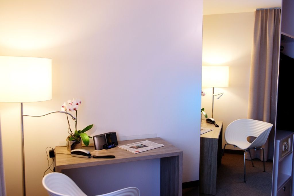 A hotel room with a desk, a chair, a lamp, a plant, and a bed.
