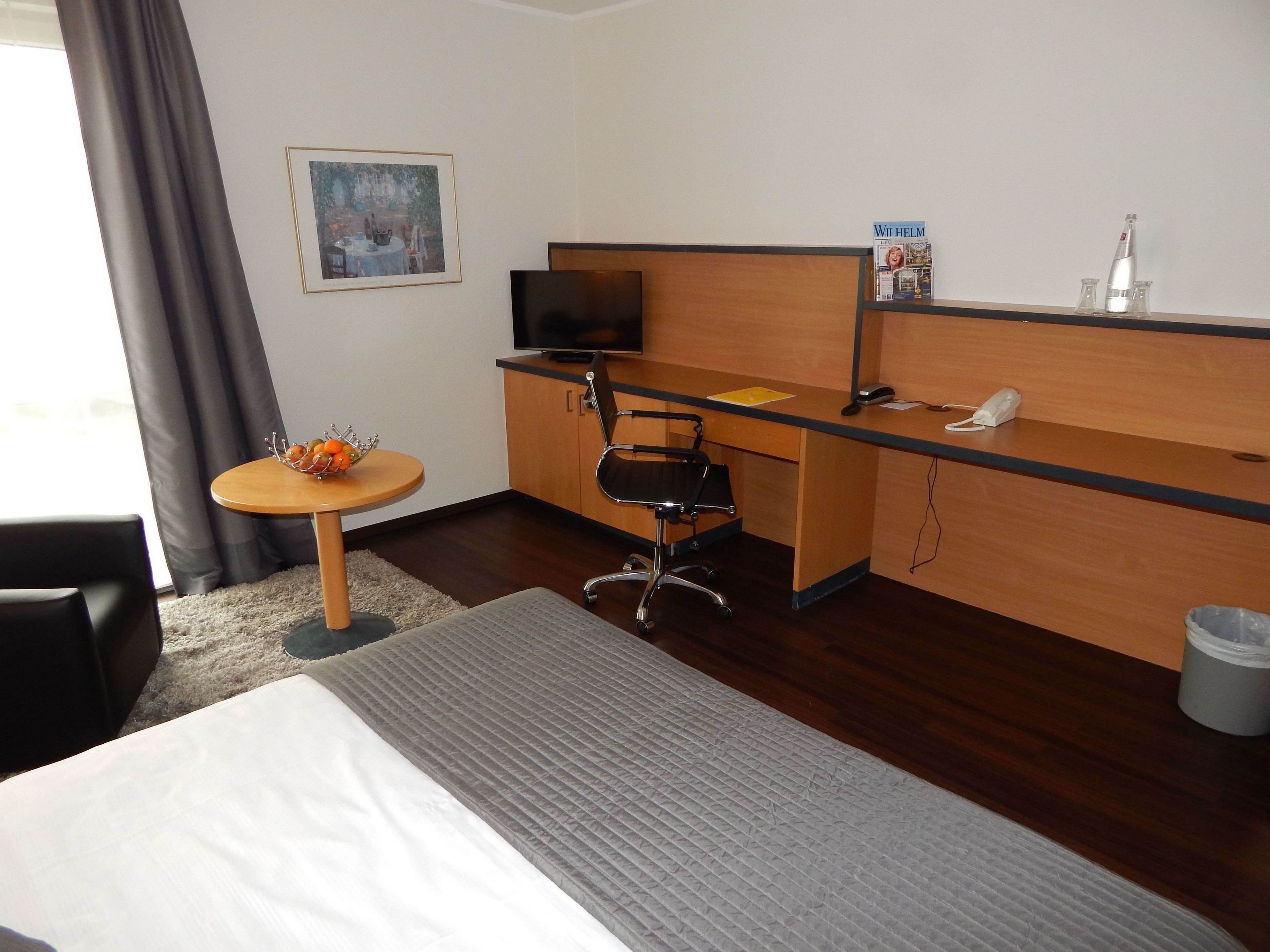 A hotel room with a bed, a desk, a chair, a TV, a small table with fruit, and a picture on the wall.