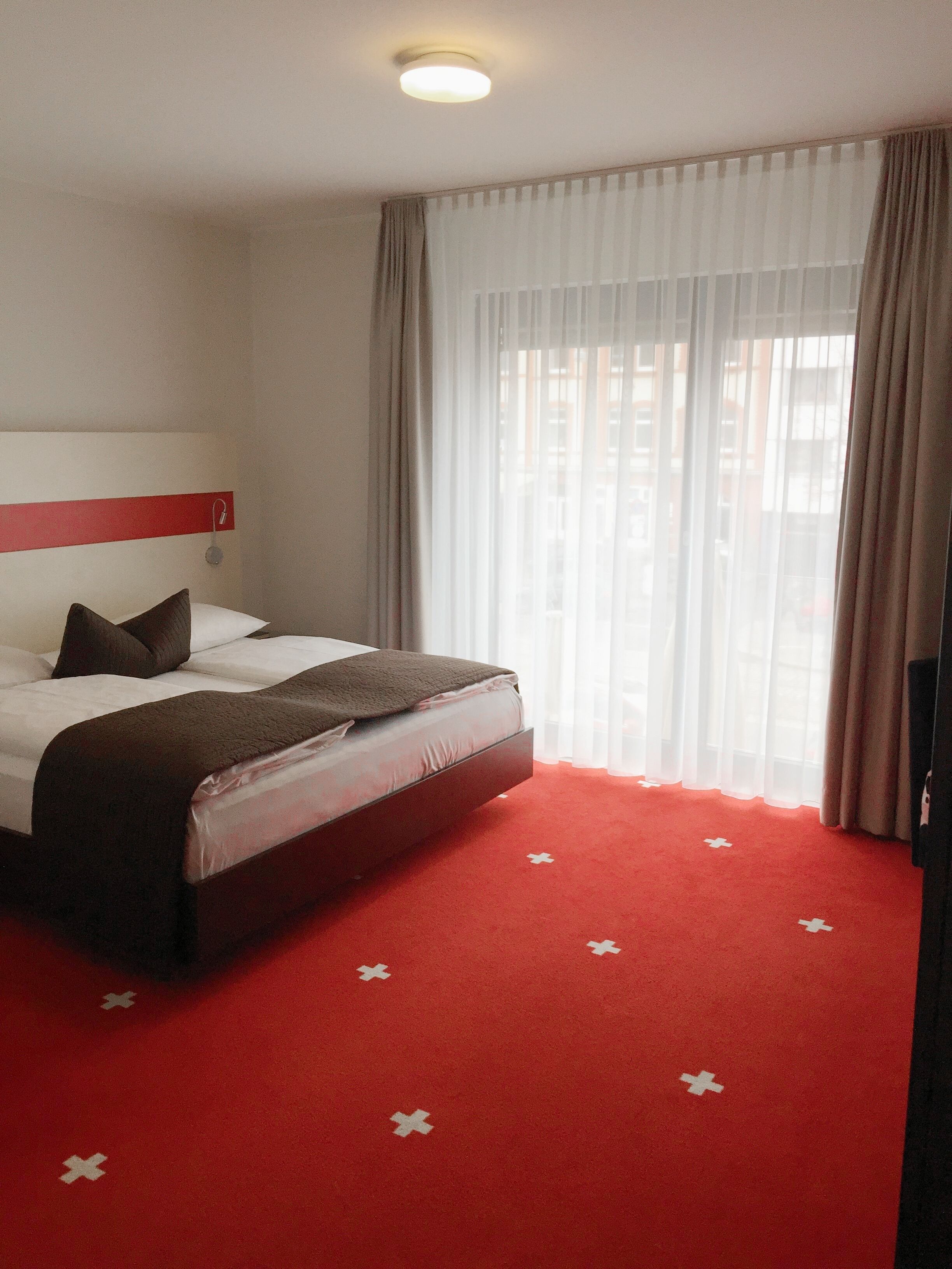 A hotel room with a bed, a red carpet with white crosses, a white headboard, and a window with sheer curtains.