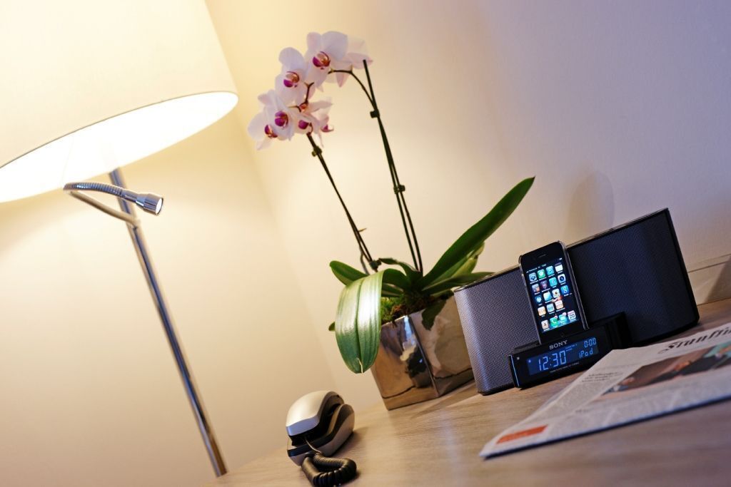 Un escritorio con una lámpara, una orquídea, un teléfono inteligente, un reloj digital y un teléfono.