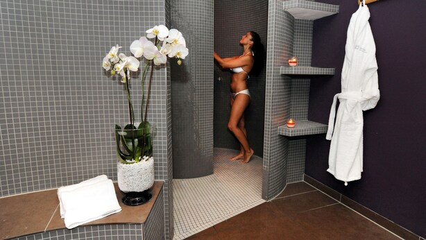 Une femme vêtue d’un bikini blanc se tient sous une douche dont les murs sont recouverts de carreaux. Une serviette blanche et une robe blanche sont suspendues à un crochet.