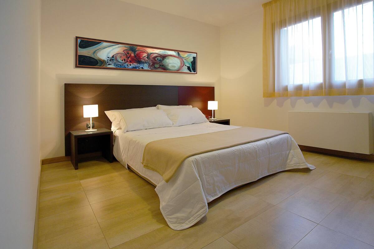 A hotel room with a large bed, two bedside tables with lamps, a painting above the bed, and a window with curtains.