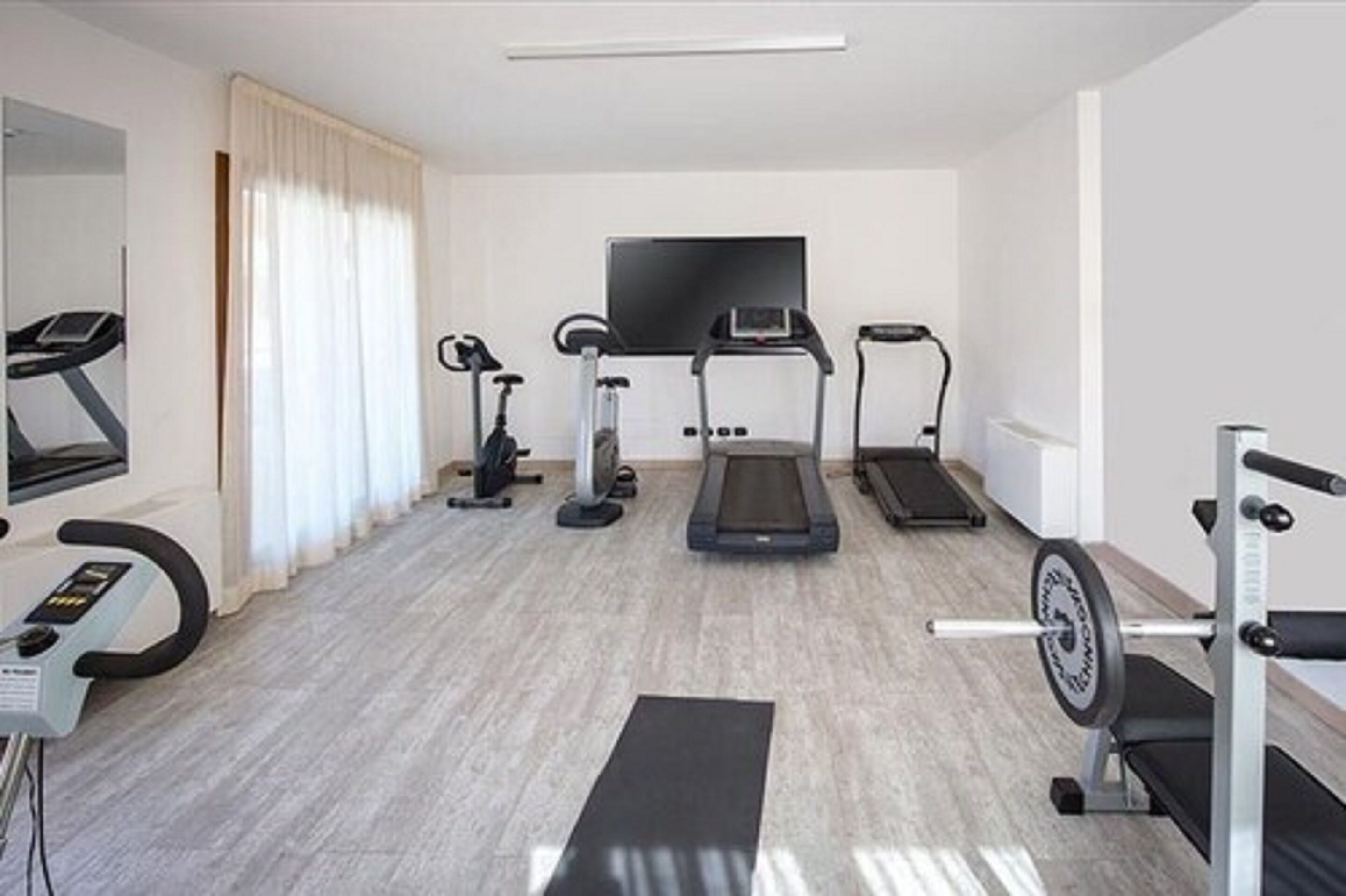 A modern hotel room with a treadmill, elliptical, stationary bike, and a TV mounted on the wall.