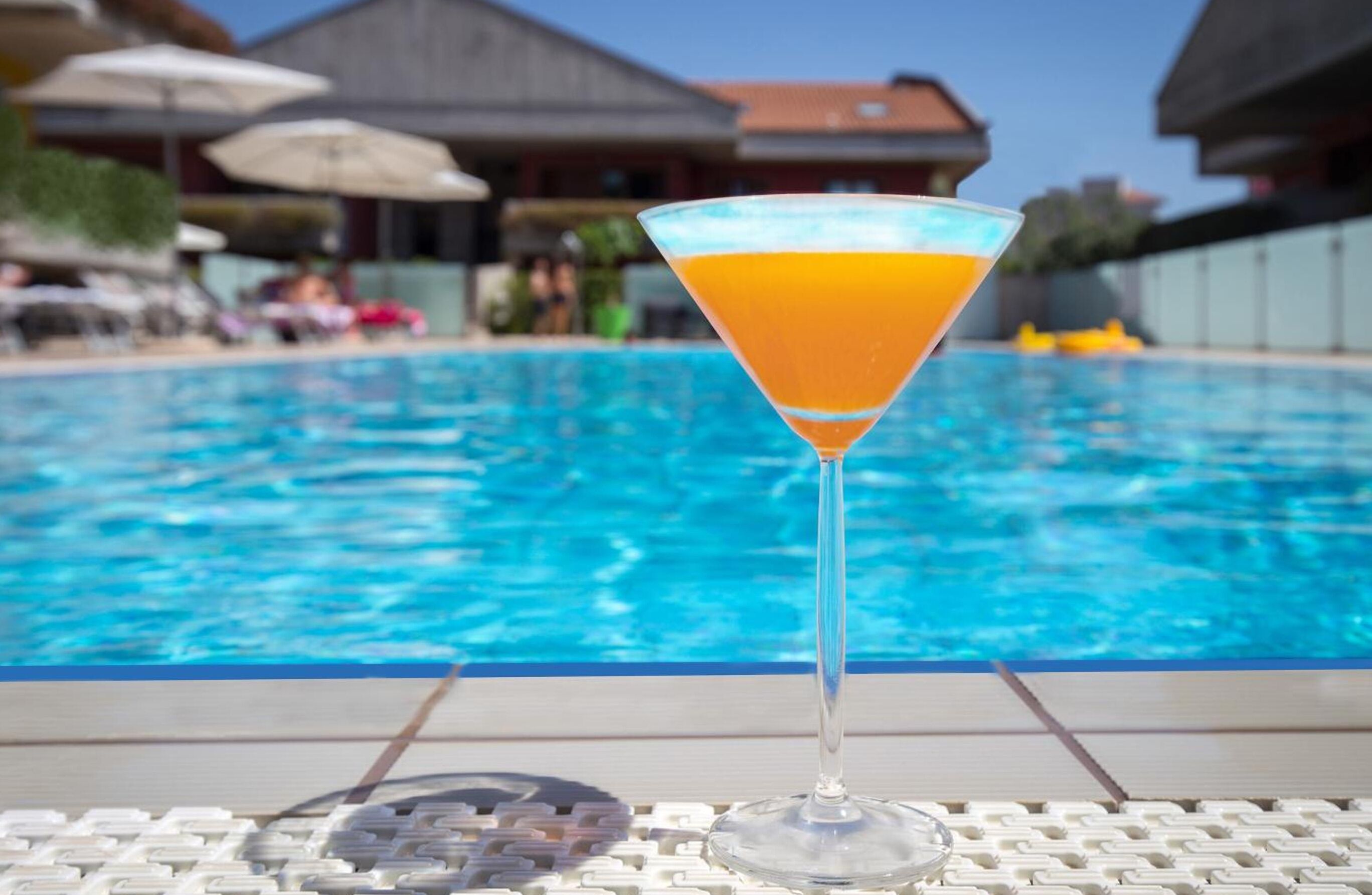 A clear glass martini glass with a yellowish-orange drink, placed on a white tiled surface by a pool.
