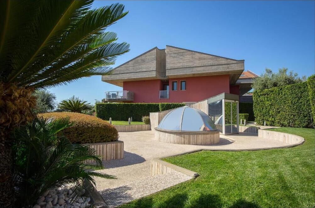 A modern house with a red facade, a large glass structure in the front, and a well-maintained garden with palm trees and hedges.