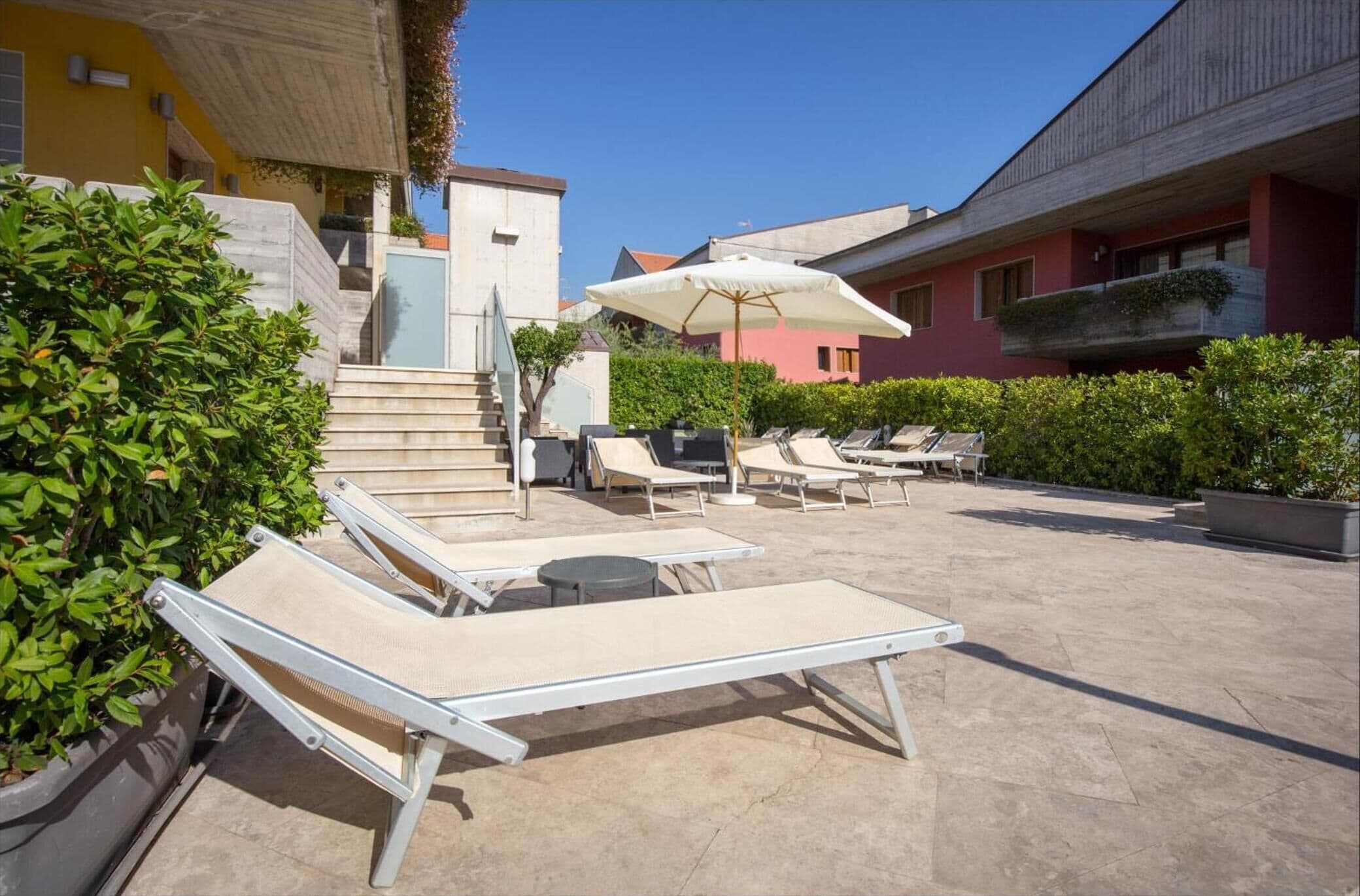A rooftop pool area with sun loungers and umbrellas, surrounded by greenery and buildings.