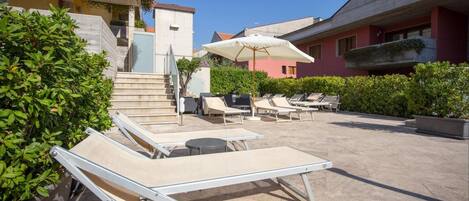 A rooftop pool area with sun loungers and umbrellas, surrounded by greenery and buildings.