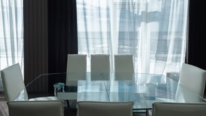 Un salón de conferencias con una mesa de vidrio y sillas blancas, ventanas amplias con cortinas translúcidas y vistas a edificios.