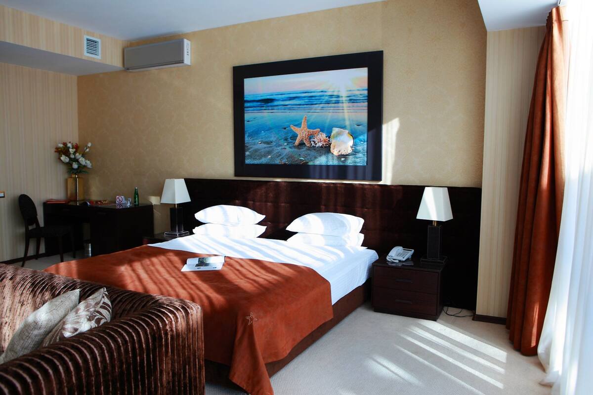A hotel room with a bed, two bedside tables, a desk, a chair, a sofa, a painting of a beach scene, and a window with curtains.
