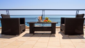 Un balcón con sillas de mimbre, una mesa con fruta y una bebida, y vista al mar.