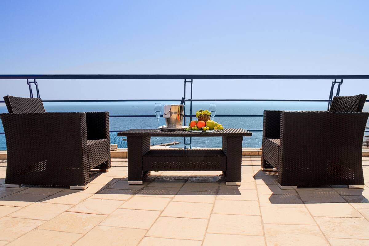 A balcony with wicker chairs, a table with fruit and a drink, and a view of the sea.