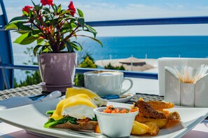 Uma bandeja de café da manhã com ovos, torradas e acompanhamentos, uma xícara de café e uma planta em vaso numa varanda com vista para o mar.