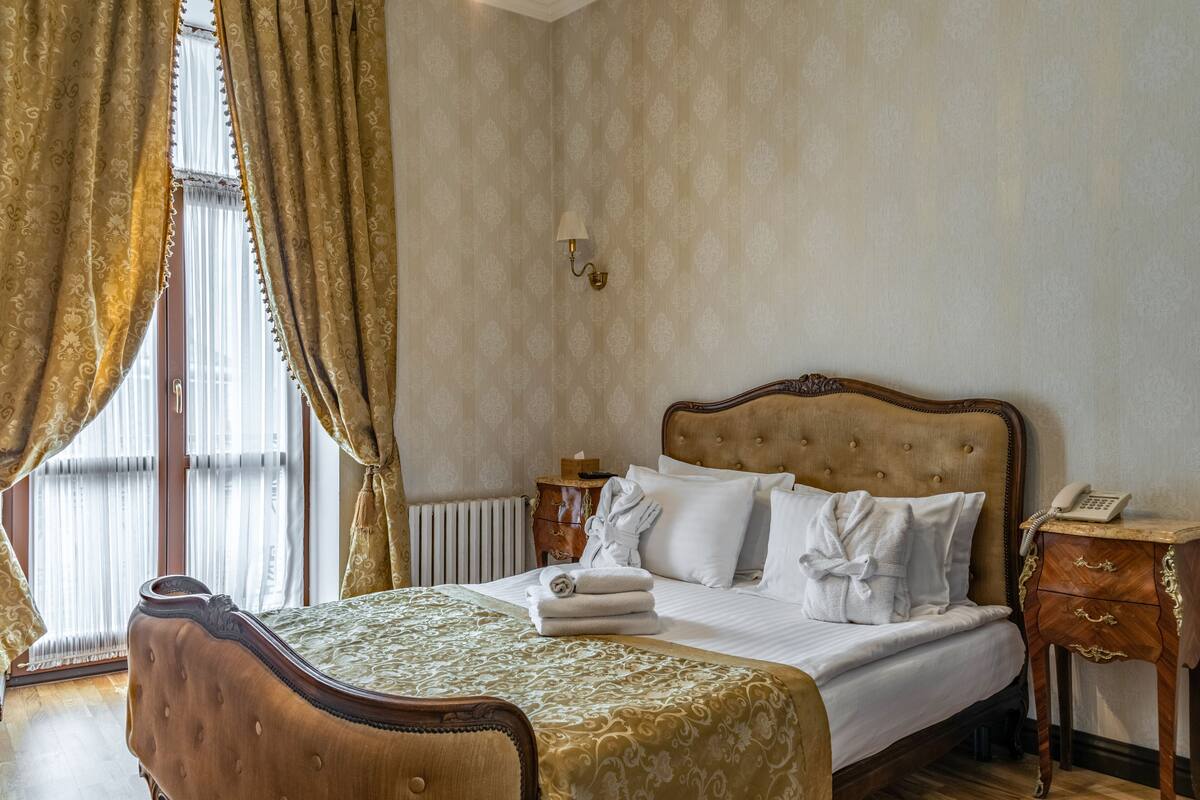 Una cama bien hecha con cabecera, una mesita de noche con lámpara y una ventana con cortinas.