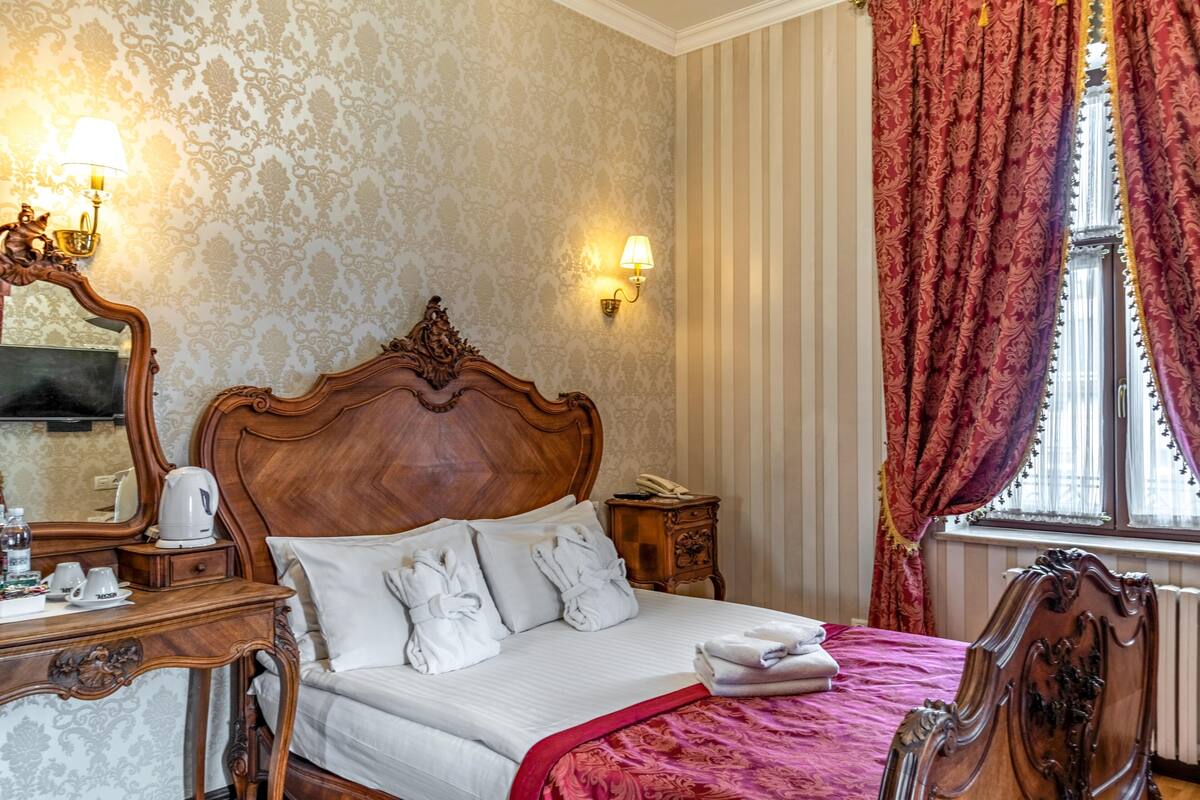 Habitación de hotel con una cama grande, cabecera de madera, mesita de noche con lámpara, espejo y ventana con cortinas.
