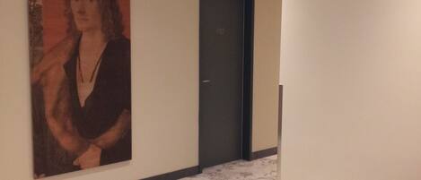 Un pasillo con un retrato grande en la pared, una puerta con la inscripción '102' y una silla roja al frente.
