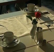 Mesa para dos con mantel blanco, un florero con una rosa roja, una taza y platillo, salero y pimentero, y una cuchara.