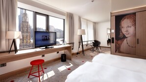 Habitación de hotel con una ventana grande, televisión, escritorio, taburete rojo, cama y un cuadro.