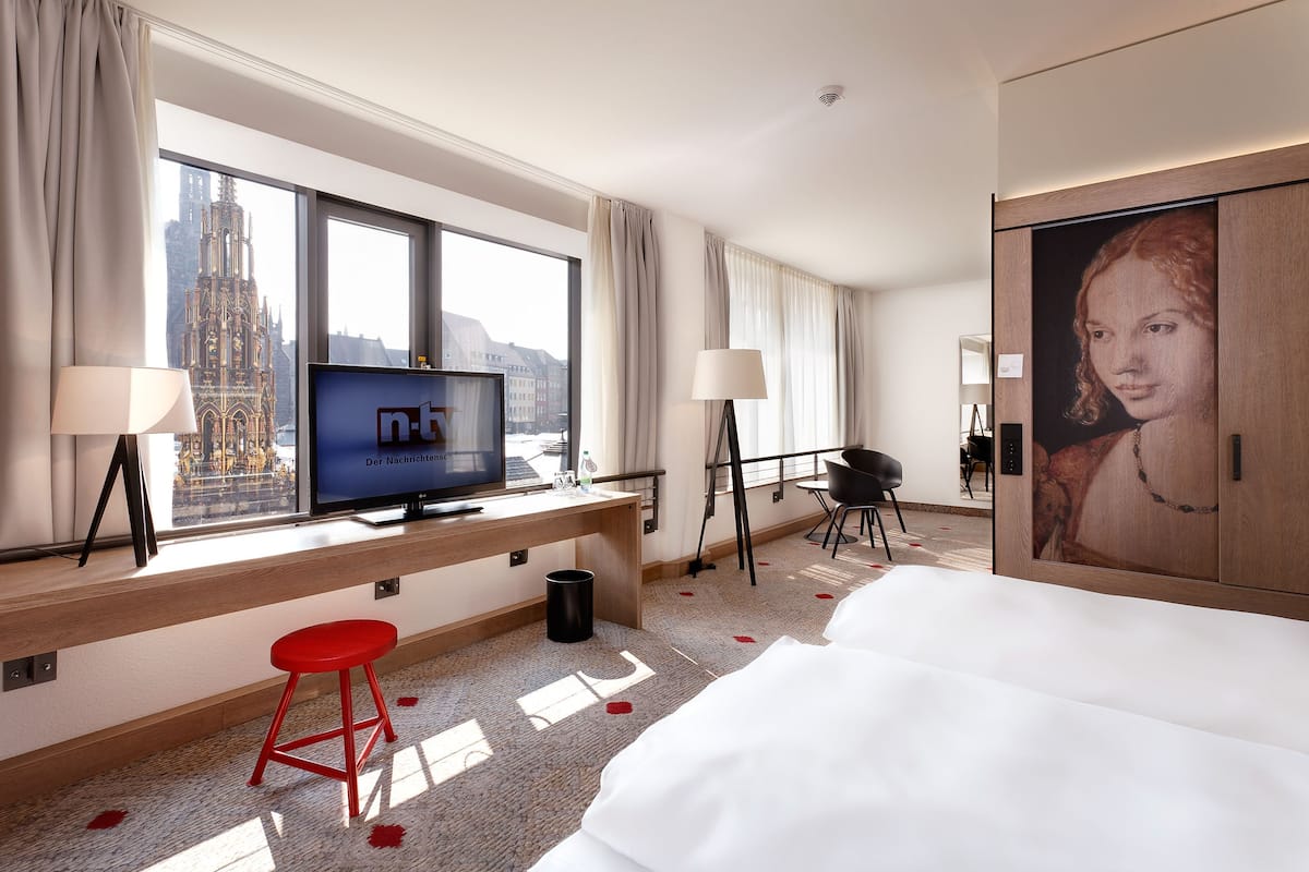 Ein Hotelzimmer mit großem Fenster, einem Fernseher, einem Schreibtisch, einem roten Hocker, einem Bett und einem Gemälde.
