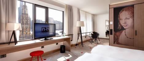 Habitación de hotel con una ventana grande, televisión, escritorio, taburete rojo, cama y un cuadro.