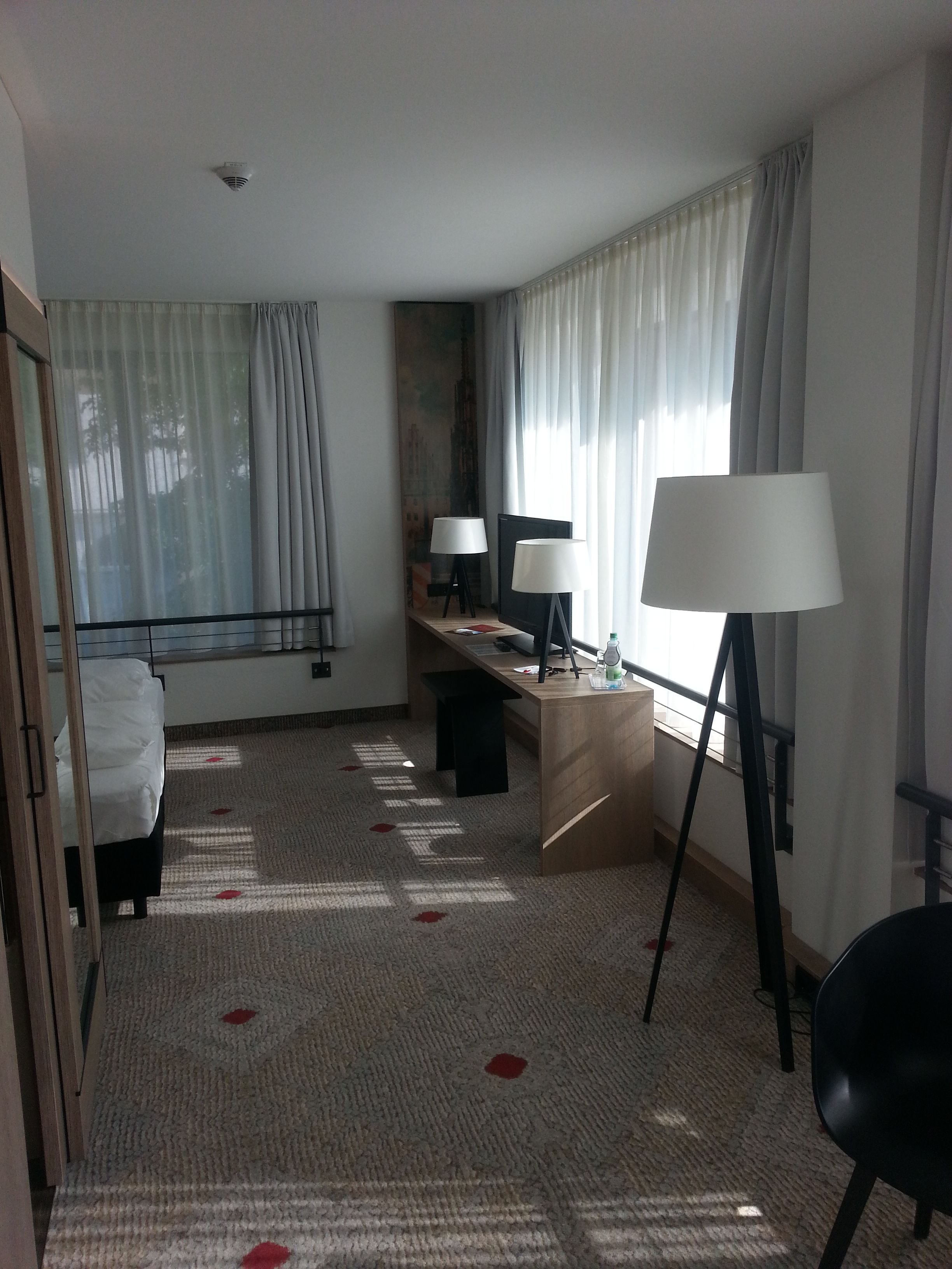 A hotel room with a bed, a desk with two lamps, a chair, and a large window with curtains.
