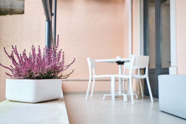 Un área de estar moderna al aire libre con una mesa y sillas blancas, una maceta con flores rosas y un edificio de pared color melocotón y grandes ventanales.