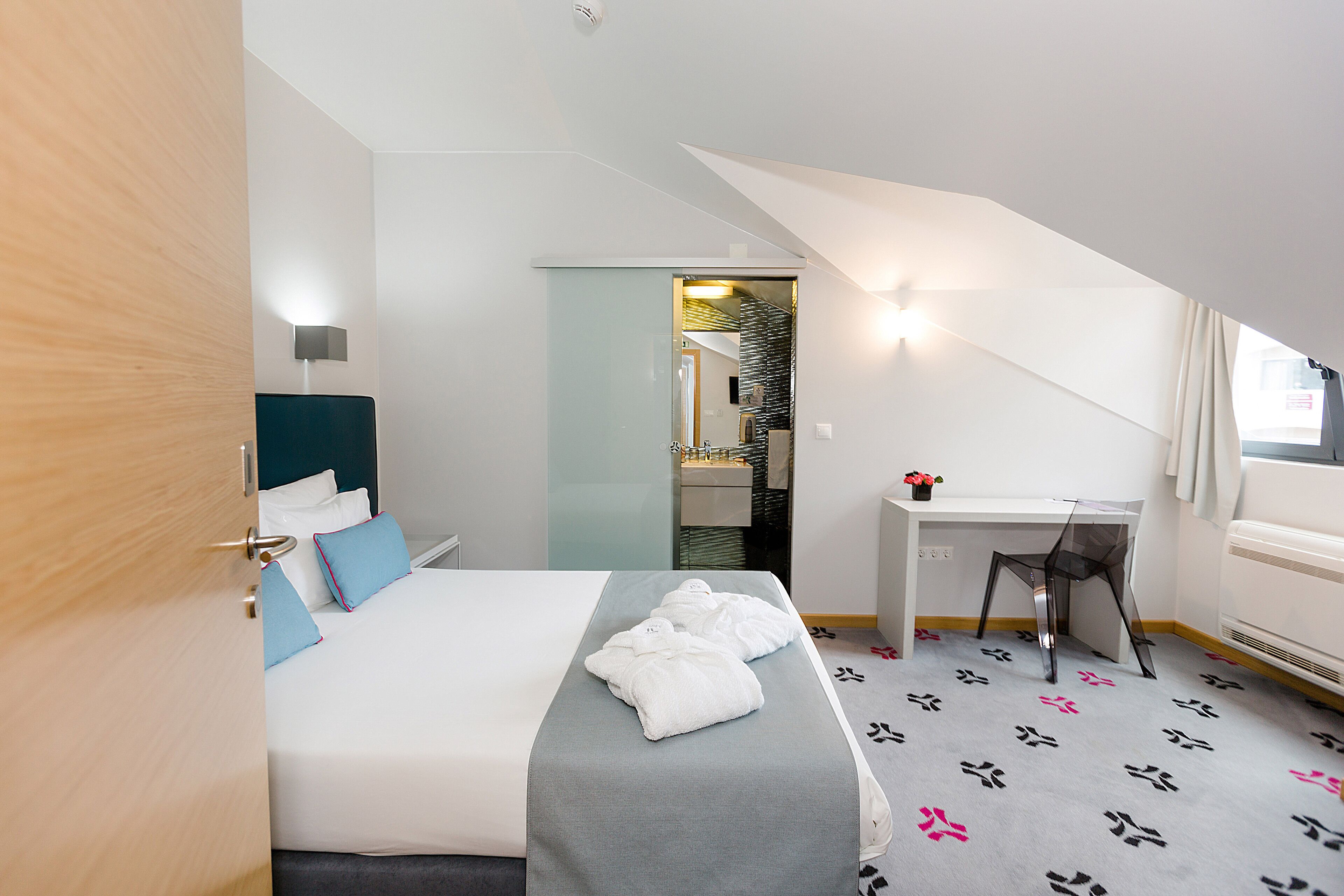 A hotel room with a bed, a desk, and a chair. There is a bathroom visible through a glass door.