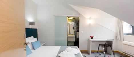 A hotel room with a bed, a desk, and a chair. There is a bathroom visible through a glass door.