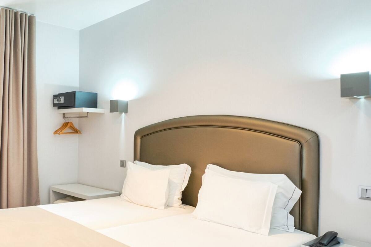Una cama doble con cabecera color beige, ropa de cama blanca, una mesita de noche con teléfono, y una lámpara de pared.
