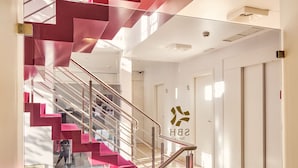A modern hotel interior with a staircase featuring pink steps and metal railings, a glass elevator, and a sign with the hotel's logo.